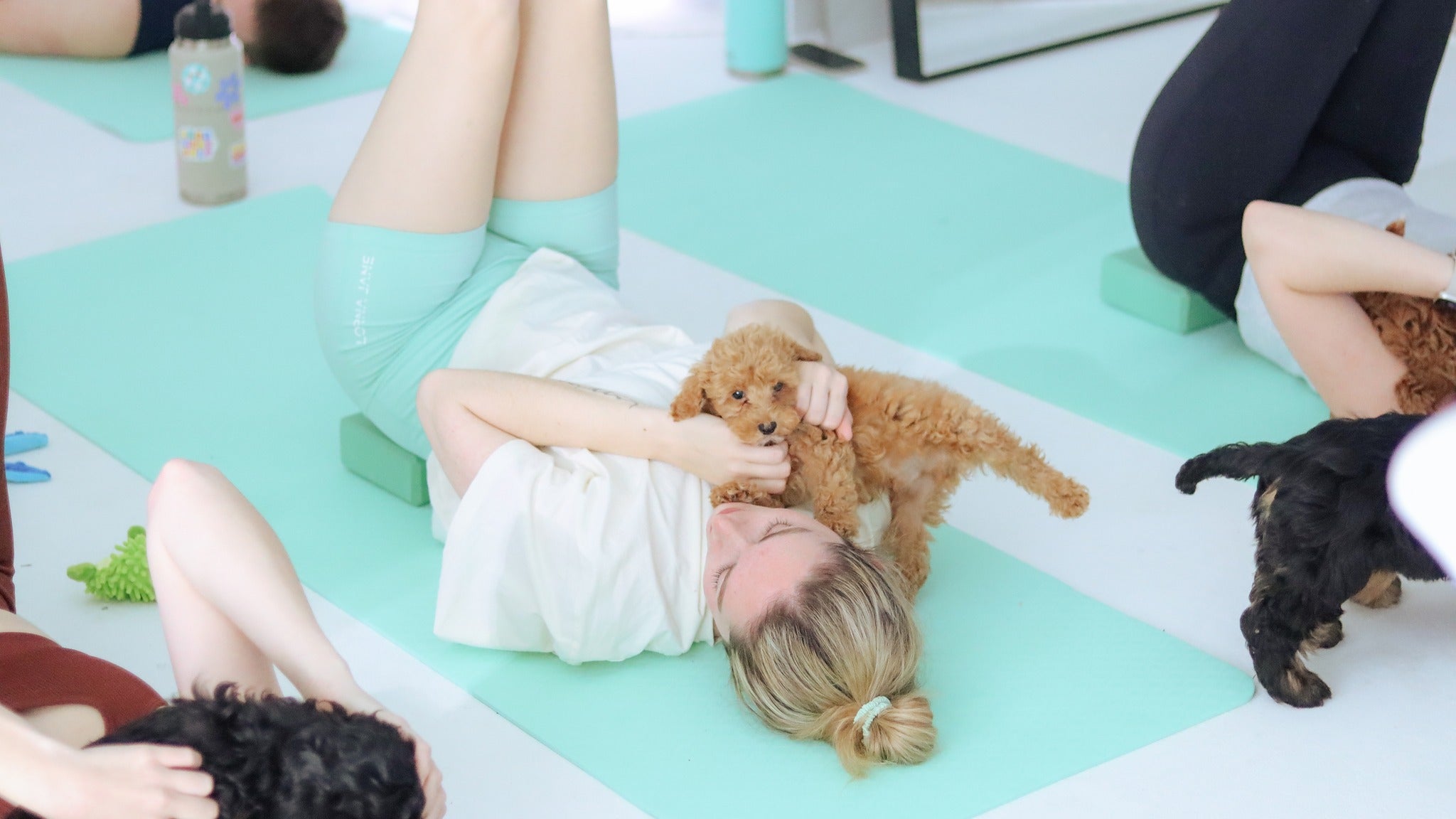 Woman lying on yoga mat during Puppy Yoga in Australia, smiling and cuddling a fluffy brown puppy